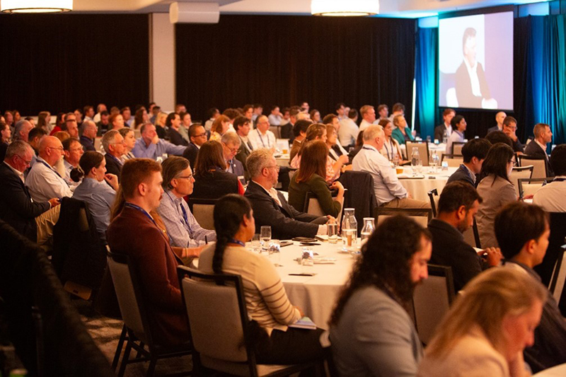 A large room of people seated at tables watching a screen presentation
