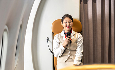 Image of a flight attendant sitting in a seat in an aircraft cabin, holding a microphone