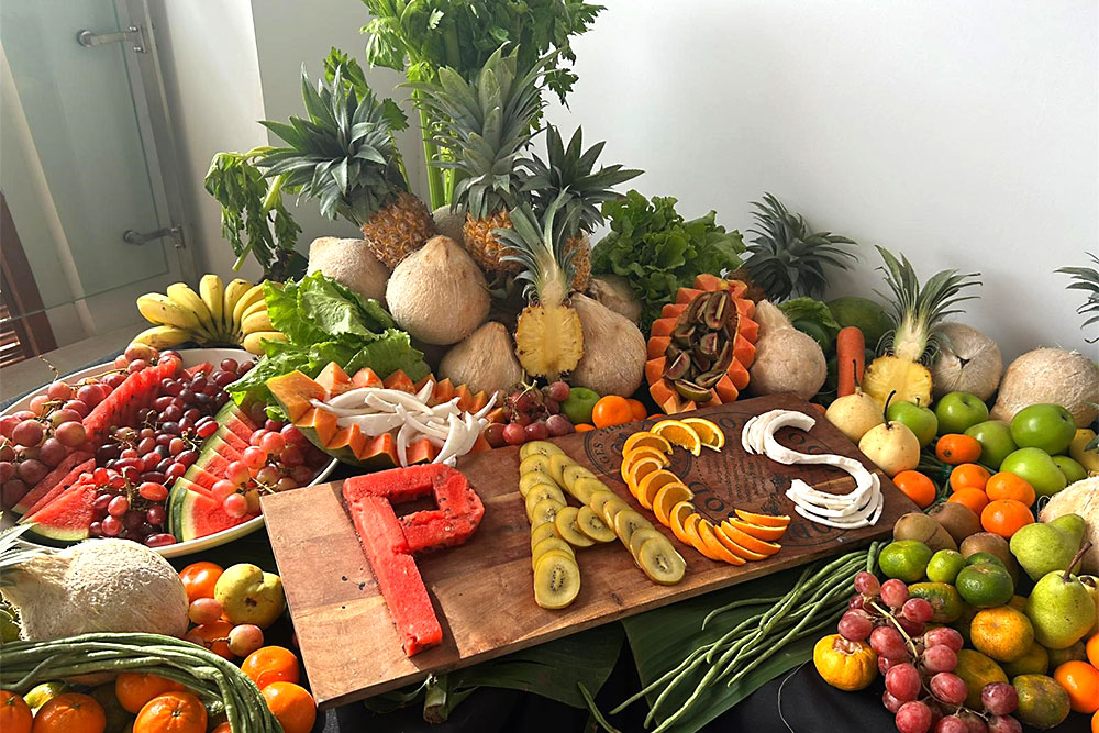 A visually striking table arrangement features a variety of whole and sliced tropical fruits. The acronym "PACS," representing the Pacific Vision for Adapted Crops and Soils, is carefully arranged on the table using slices of watermelon, kiwifruit, orange, and coconut.