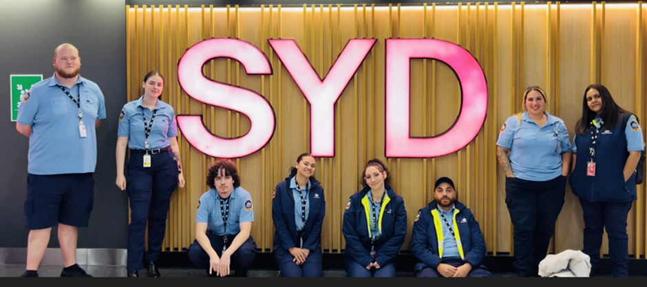8 people sitting and standing in front of a wall with a sign displaying the letters "SYD"