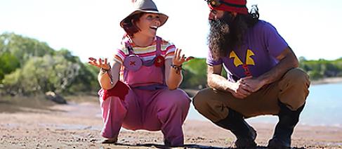 Image of two people crouching down on sand, with trees and some water in the background