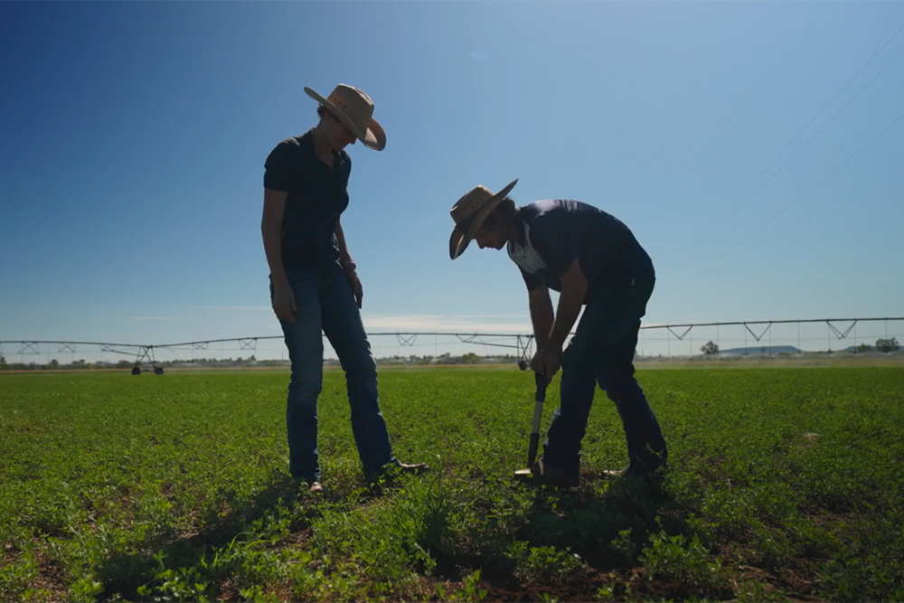 Image of two people in a green field with an irrigation machine in the background, one person is digging with a small shovel as the other observes