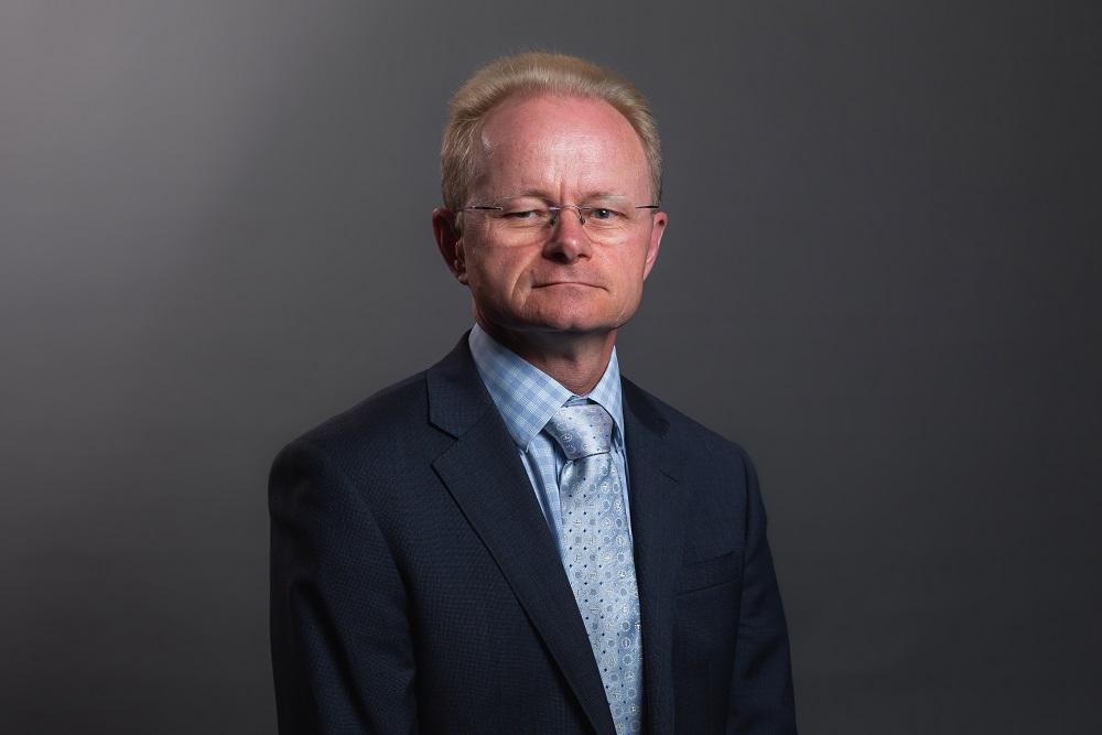 Formal portrait photo of Dr Mark Schipp in a dark suit with a blue shirt and tie.