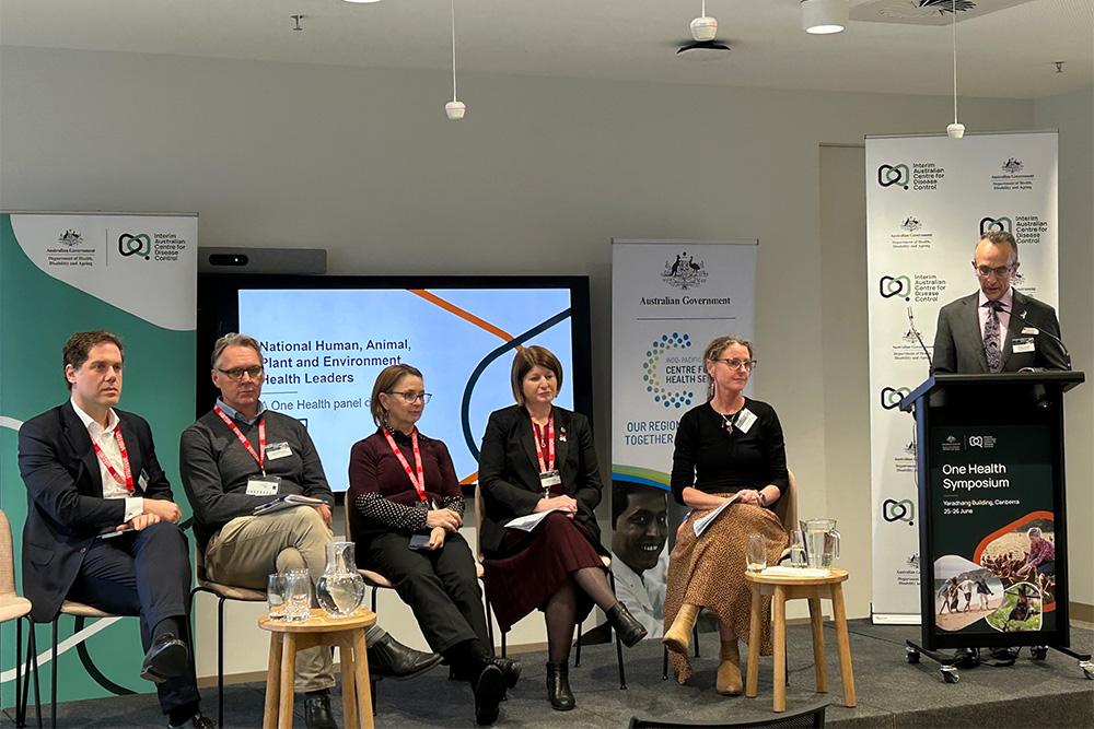 Image of 5 people sitting in front of an audience and one person standing at a podium