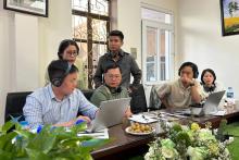 6 Vienatmese people gathered around a long desk containing computers and refreshments, in deep discussion.